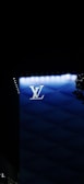 A modern architectural structure features a prominent illuminated brand logo 'LV' against a backdrop of gradient blue panels. Small lights outline the edge of the building, while a shadowy tree partially frames the right side.
