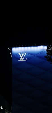 A modern architectural structure features a prominent illuminated brand logo 'LV' against a backdrop of gradient blue panels. Small lights outline the edge of the building, while a shadowy tree partially frames the right side.