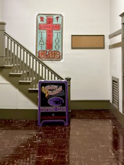 A hallway corner with a staircase on the left and two large signs for a tattoo club. The larger poster is vertically oriented, featuring an intricate design with the text 'Violent Delights Tattoo Club' in a stylized font. Below it is a smaller, standing sign with similar branding. The walls are painted white, and the staircase is light gray with matching handrails. The floor is dark brown, reflecting light from the walls.