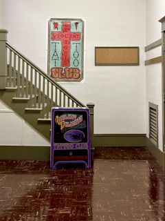 A hallway corner with a staircase on the left and two large signs for a tattoo club. The larger poster is vertically oriented, featuring an intricate design with the text 'Violent Delights Tattoo Club' in a stylized font. Below it is a smaller, standing sign with similar branding. The walls are painted white, and the staircase is light gray with matching handrails. The floor is dark brown, reflecting light from the walls.