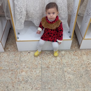 A child wearing a red dress with black spots and a furry collar is sitting on a white display shelf. The child has white leggings and yellow shoes. The background includes a patterned curtain and a tiled floor.