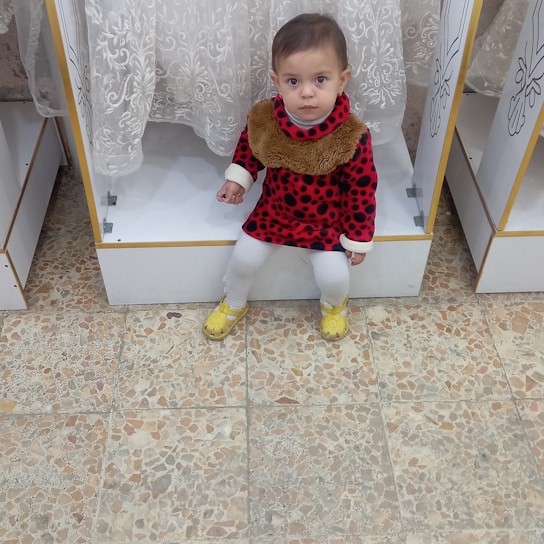 A child wearing a red dress with black spots and a furry collar is sitting on a white display shelf. The child has white leggings and yellow shoes. The background includes a patterned curtain and a tiled floor.