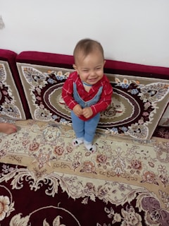 A small child wearing a red shirt and blue overalls is standing on an ornately patterned carpet. The child is smiling and looks happy, positioned in front of a patterned sofa or cushion.