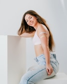 A young woman with long brown hair and a smile poses in a relaxed manner. She is leaning against a white surface and is wearing a white top and light blue jeans. Her arms have decorative temporary tattoos.