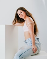 A young woman with long brown hair and a smile poses in a relaxed manner. She is leaning against a white surface and is wearing a white top and light blue jeans. Her arms have decorative temporary tattoos.