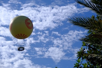 A large yellow hot air balloon featuring a logo or emblem is floating in a sky filled with scattered white clouds. To the right, the silhouette of palm tree leaves is visible, adding a natural element to the scene.