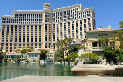A large multi-story building with numerous windows, known for its luxurious design, stands next to a body of water surrounded by greenery. The structure has a prominent and elegant architectural style. In front, there is a green area with trees, shrubs, and a brand-name retail shop with 'Louis Vuitton' signage.