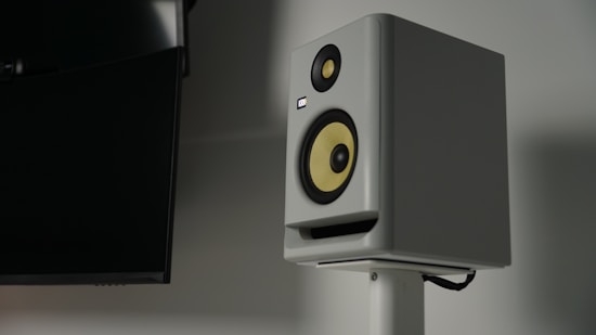A modern studio monitor speaker with a white casing and a yellow speaker cone mounted on a stand. The room is dimly lit, and there is a large, black, flat surface visible on the left side, possibly a television or a computer monitor.