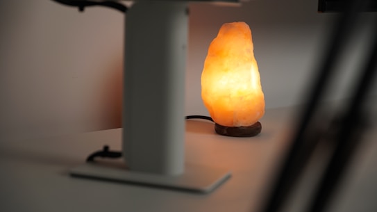 A warm, glowing salt lamp sits on a flat surface, casting a soft, ambient light. It's next to a metallic structure, likely a part of a stand or table, partially visible in the foreground.