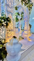 An elegantly decorated setting with intricate white archways adorned with floral arrangements and hanging greenery. Soft lighting enhances the detailed carvings, creating a serene and sophisticated atmosphere. A white rug and two ornate white vases are positioned prominently.