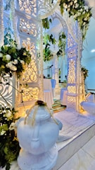 An elegantly decorated setting with intricate white archways adorned with floral arrangements and hanging greenery. Soft lighting enhances the detailed carvings, creating a serene and sophisticated atmosphere. A white rug and two ornate white vases are positioned prominently.