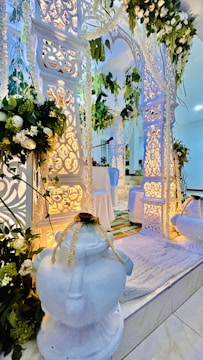 An elegantly decorated setting with intricate white archways adorned with floral arrangements and hanging greenery. Soft lighting enhances the detailed carvings, creating a serene and sophisticated atmosphere. A white rug and two ornate white vases are positioned prominently.