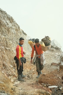 Two individuals are on a rough, rocky path. They appear to be carrying heavy loads of sulfur in baskets over their shoulders. The landscape around them is earthy and rugged, suggesting a mining area.