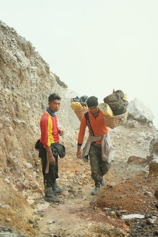 Two individuals are on a rough, rocky path. They appear to be carrying heavy loads of sulfur in baskets over their shoulders. The landscape around them is earthy and rugged, suggesting a mining area.