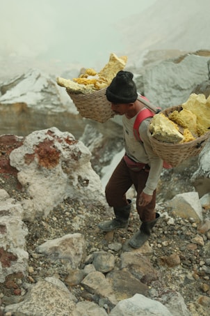 A person is carrying two large baskets filled with yellow sulfur on their back while navigating a rocky terrain. The scene is set in a mountainous area with visible stones and rugged surfaces. The individual is wearing a black beanie, a long-sleeve shirt, pants, and boots.