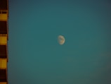A large moon is visible in the sky against a teal background. The moon is illuminated, showing clear surface details. On the left, the edge of a building with visible balconies is partially shown in shadow.