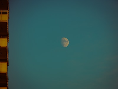 A large moon is visible in the sky against a teal background. The moon is illuminated, showing clear surface details. On the left, the edge of a building with visible balconies is partially shown in shadow.