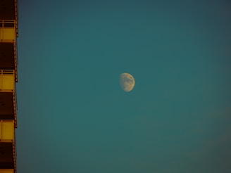 A large moon is visible in the sky against a teal background. The moon is illuminated, showing clear surface details. On the left, the edge of a building with visible balconies is partially shown in shadow.