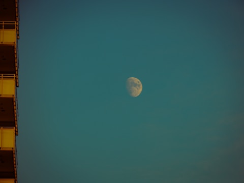 A large moon is visible in the sky against a teal background. The moon is illuminated, showing clear surface details. On the left, the edge of a building with visible balconies is partially shown in shadow.