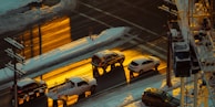 An urban street scene during winter, showcasing vehicles driving on a snow-lined road. The road is bathed in a warm golden light, likely from the setting sun, creating a stark contrast with the surrounding snow. There are several cars moving in both directions, and a structure is seen on the right side.