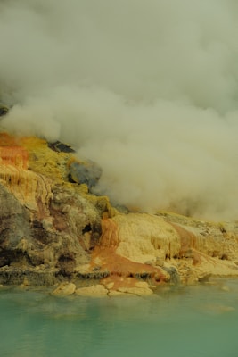 A landscape featuring a geothermal area with colorful mineral deposits and steam rising from the ground. The scene includes a mix of colors such as yellow, red, brown, and black minerals, alongside turquoise water in the foreground. Thick clouds of steam obscure parts of the scene, creating a surreal and somewhat mystical atmosphere.