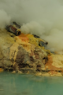 A geothermal landscape with fumaroles emitting steam, where mineral deposits create vibrant hues of yellow, orange, and brown on the rocky terrain. The foreground features a pale turquoise pool of water.