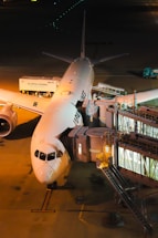 A commercial airplane is parked at an airport gate, connected to a jet bridge for passenger boarding. The plane has visible airline branding along its fuselage. The scene is illuminated by artificial lighting, casting a warm glow on the tarmac. Airport service vehicles, including catering trucks, are positioned nearby, indicating preparations for departure.