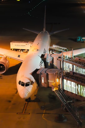 A commercial airplane is parked at an airport gate, connected to a jet bridge for passenger boarding. The plane has visible airline branding along its fuselage. The scene is illuminated by artificial lighting, casting a warm glow on the tarmac. Airport service vehicles, including catering trucks, are positioned nearby, indicating preparations for departure.