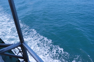 Waves created by a boat moving through the ocean are visible, with foam forming along the edge. Part of the boat's railing is visible on the left, indicating motion through water. The sea is a deep blue color, with a clear sky reflection.