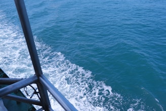 Waves created by a boat moving through the ocean are visible, with foam forming along the edge. Part of the boat's railing is visible on the left, indicating motion through water. The sea is a deep blue color, with a clear sky reflection.