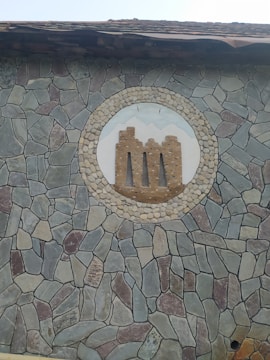 A stone wall is decorated with an intricate pattern of multicolored stones, including grey, brown, and beige tones. At the center of the wall is a circular mosaic depicting a stylized castle-like structure in brick, surrounded by a border of smaller stones. Above the mosaic is a wooden roof edge.