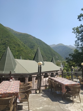 A scenic outdoor dining area with tables and chairs arranged on a terrace overlooking a mountainous landscape. The rooftops feature pointed, conical structures with green tiles, against a backdrop of lush green hills and a clear blue sky. There is a railing along the terrace and the space is shaded by a few small trees.