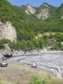 A scenic mountain landscape with lush green vegetation covering the slopes and rocky formations. The foreground features a dry riverbed with large rocks and a couple of tables with chairs set up on the riverbank.
