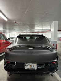 A sleek, matte grey Aston Martin car parked in an indoor parking garage. The car has a prominent Aston Martin logo on the back with an exhaust on either side. It is parked next to a red vehicle, with concrete pillars and fluorescent lighting visible.