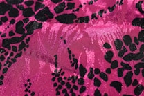 A bright pink fabric with an abstract black leopard print pattern. The material has a shiny texture, with light reflecting off the surface, highlighting the intricate design.