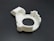 A white, circular 3D-printed plastic object with a cutout resembling a gear or mechanical part. It has various geometric shapes and indents on its surface, placed on a dark background.