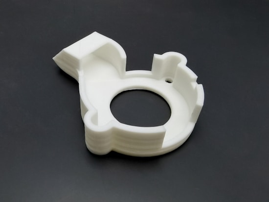 A white, circular 3D-printed plastic object with a cutout resembling a gear or mechanical part. It has various geometric shapes and indents on its surface, placed on a dark background.