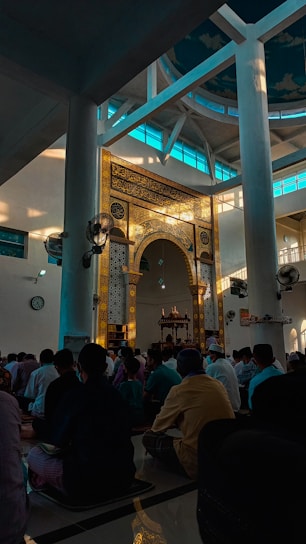 A group of people seated inside a mosque, with intricate Arabic calligraphy and decorative patterns adorning a central arch. Sunlight filters through large windows, casting a serene glow over the room.