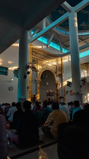 A group of people seated inside a mosque, with intricate Arabic calligraphy and decorative patterns adorning a central arch. Sunlight filters through large windows, casting a serene glow over the room.