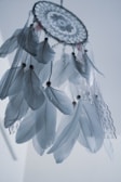 An intricately designed dreamcatcher adorned with white feathers and lace ribbons. The dreamcatcher features a web-like center with beads scattered across the hanging feathers which appear soft and delicate. The background is a soft bluish-white, providing a serene contrast to the dreamcatcher.