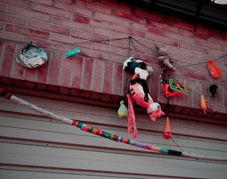 Various colorful objects including a stuffed toy tiger, a small plastic fish-shaped container, a small cone, and multiple other hanging items are attached to strings against a brick wall. There is also a colorfully painted stick or pole running horizontally at the bottom of the image.