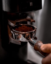 A hand is holding a coffee portafilter filled with freshly ground coffee, positioned underneath an espresso machine ready for brewing. The focus is on the rich, dark coffee grounds and the metallic sheen of the portafilter.
