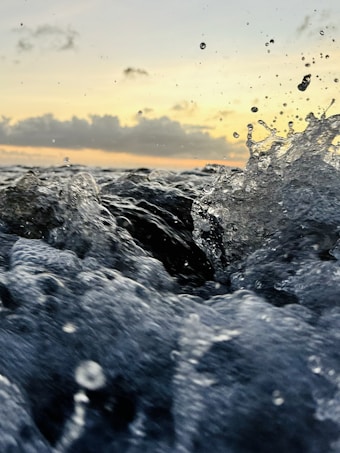 Waves crashing energetically against each other with droplets of water captured mid-air. The background shows a serene sunset with soft clouds covering parts of the horizon.