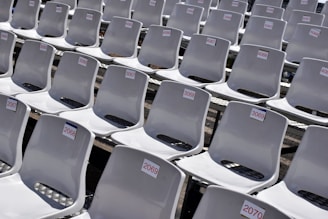 Rows of empty white plastic seats, each labeled with numbered tags, are neatly arranged in a stadium-like setting. The seats are aligned in perfectly straight rows on metal bleachers, creating a sense of order and uniformity.