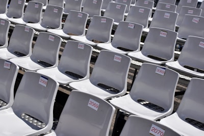Rows of empty white plastic seats, each labeled with numbered tags, are neatly arranged in a stadium-like setting. The seats are aligned in perfectly straight rows on metal bleachers, creating a sense of order and uniformity.