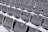 Rows of empty white plastic seats, each labeled with numbered tags, are neatly arranged in a stadium-like setting. The seats are aligned in perfectly straight rows on metal bleachers, creating a sense of order and uniformity.