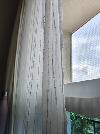 A sheer white curtain with subtle decorative patterns hangs next to a window. Beyond the window, there is a view of a clear sky and some greenery. The window includes a mesh screen, allowing light to filter softly into the room. To the side, a metallic bottle is visible, partially in shadow.
