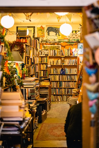 A cozy, cluttered bookstore filled with tightly packed shelves of books. Warm lighting highlights an eclectic assortment of decorations, including posters and hanging lights. The space has a lived-in, inviting atmosphere, suggesting a place for leisurely browsing.