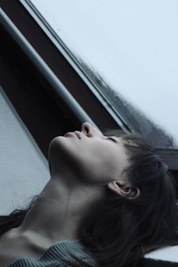 A person is seen in a pensive pose, with their head tilted back against a window. The scene appears introspective with soft lighting and a gentle focus on the contours of their face.