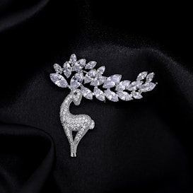 A silver brooch designed in the shape of a stylized deer, adorned with sparkling gemstones that resemble a branch with leaves. The background is a smooth, black fabric that enhances the elegance of the jewelry piece.